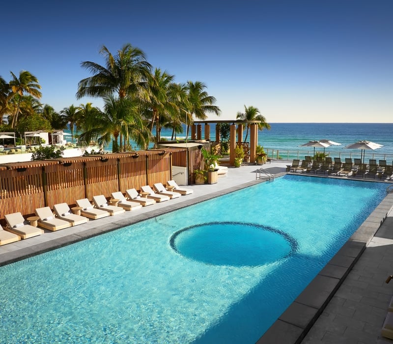 A long, light-blue swimming pool features a circular area, bordered by lounge chairs and tropical palm trees, overlooking the ocean under a clear sky.