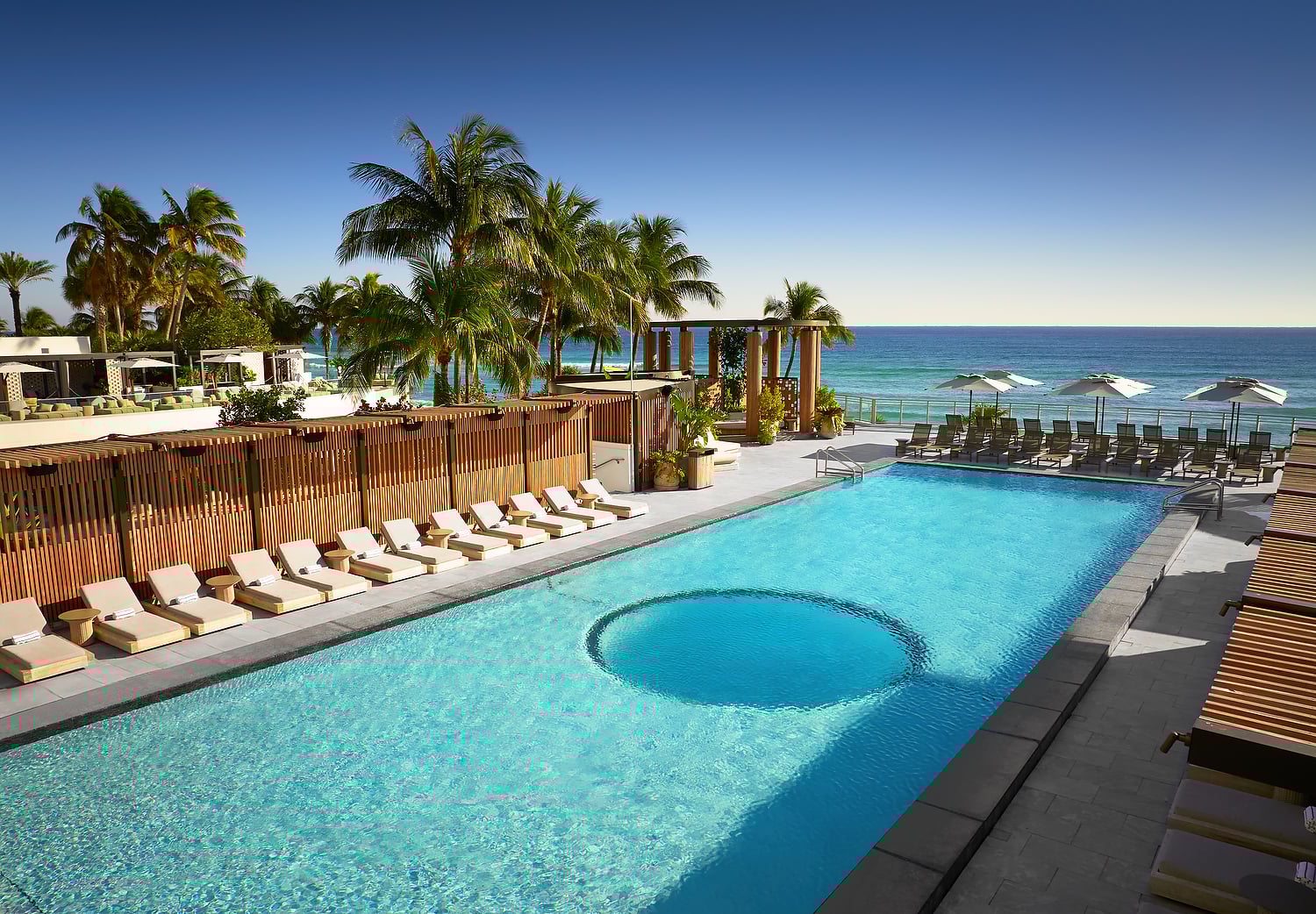 A long, light-blue swimming pool features a circular area, bordered by lounge chairs and tropical palm trees, overlooking the ocean under a clear sky.