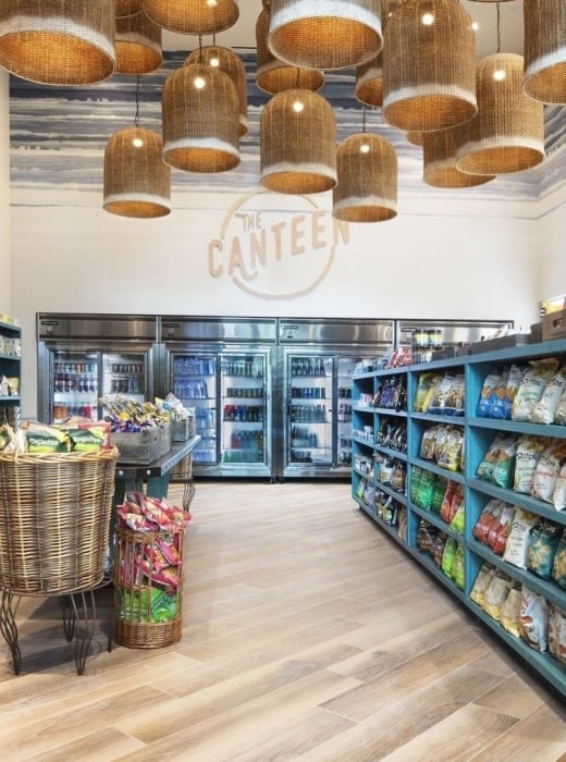 A modern canteen interior displays snacks and drinks on teal shelving, with many woven pendant lights. "THE CANTEEN" is painted on the back wall.
Text: THE CANTEEN, lolli pop, TROUT, FALLEN PRETZEL, Chips.