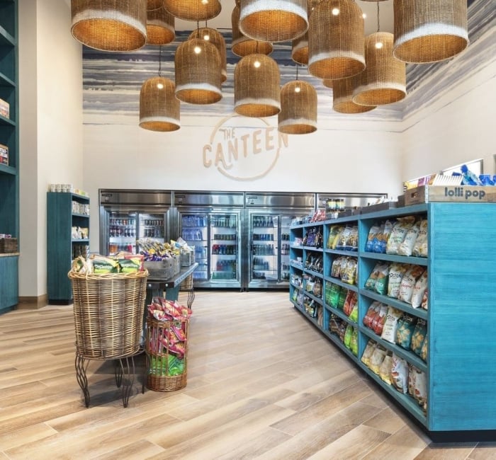 A modern canteen interior displays snacks and drinks on teal shelving, with many woven pendant lights. "THE CANTEEN" is painted on the back wall.
Text: THE CANTEEN, lolli pop, TROUT, FALLEN PRETZEL, Chips.