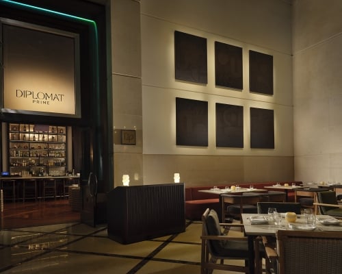 A warmly lit restaurant entrance with a backlit sign reading "DIPLOMAT PRIME" and a "DP" plaque, opens to a full bar and a dining room with set tables and six dark abstract artworks.