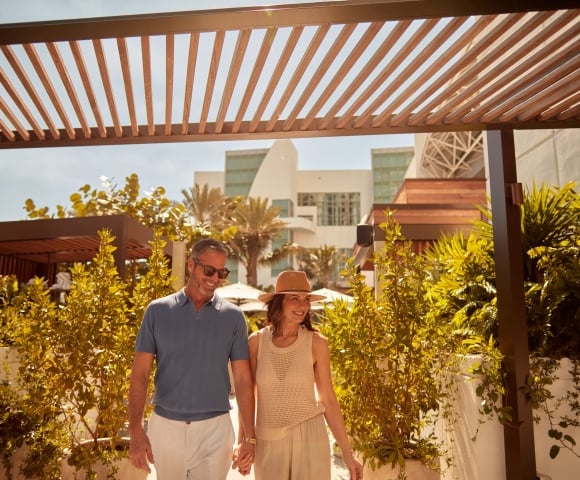 A smiling couple walks hand-in-hand under a slatted wooden pergola. Lush green foliage surrounds them, with a modern white building and palm trees visible behind them.