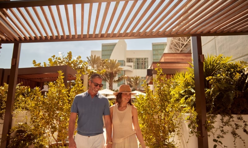 A smiling couple walks hand-in-hand under a slatted wooden pergola. Lush green foliage surrounds them, with a modern white building and palm trees visible behind them.
