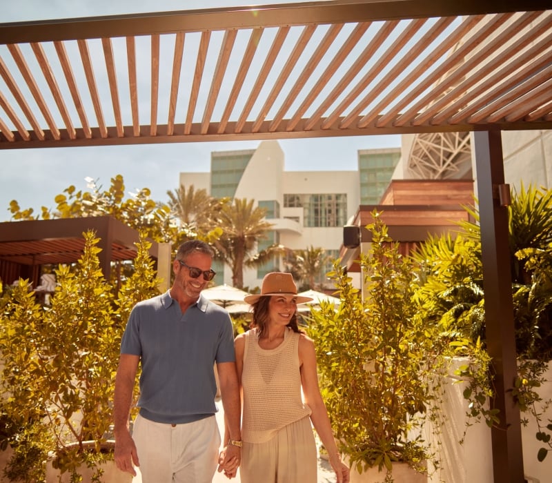 A smiling couple walks hand-in-hand under a slatted wooden pergola. Lush green foliage surrounds them, with a modern white building and palm trees visible behind them.