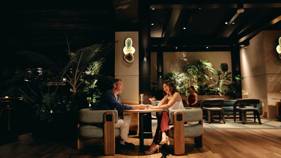 A smiling man and woman sit at a table, holding hands and talking over cocktails in a dimly lit, upscale lounge with wood floors and lush plants.