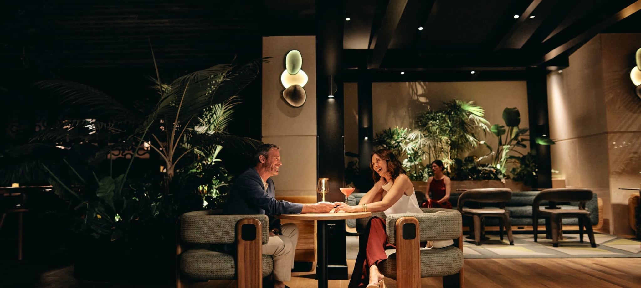 A smiling man and woman sit at a table, holding hands and talking over cocktails in a dimly lit, upscale lounge with wood floors and lush plants.