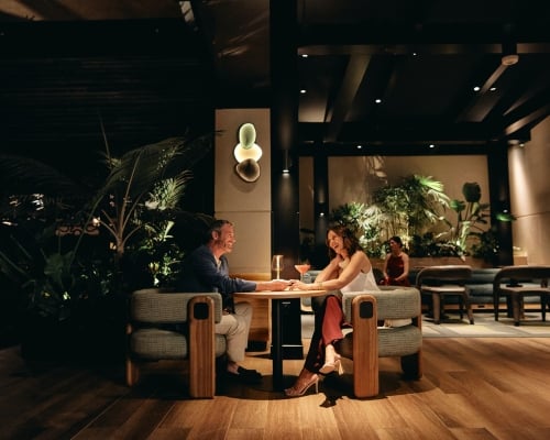 A smiling man and woman sit at a table, holding hands and talking over cocktails in a dimly lit, upscale lounge with wood floors and lush plants.
