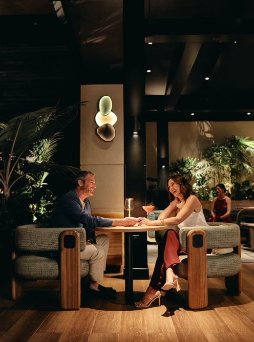A smiling man and woman sit at a table, holding hands and talking over cocktails in a dimly lit, upscale lounge with wood floors and lush plants.