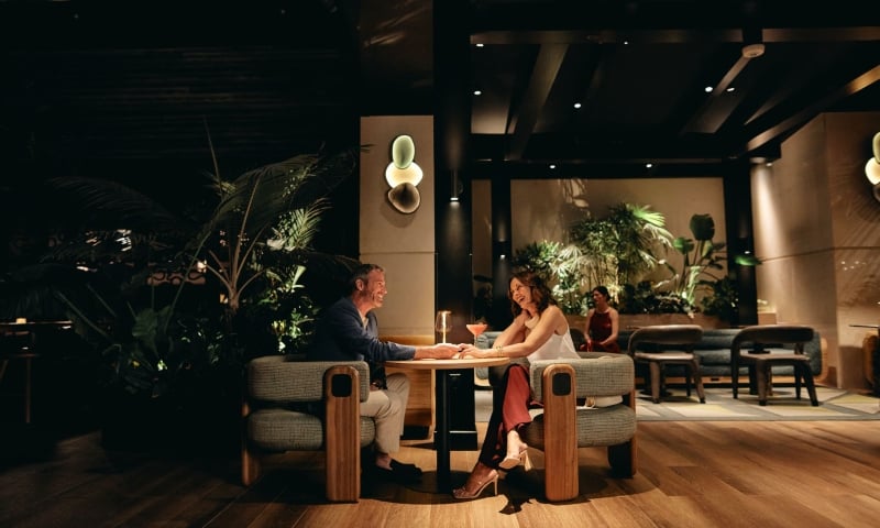 A smiling man and woman sit at a table, holding hands and talking over cocktails in a dimly lit, upscale lounge with wood floors and lush plants.