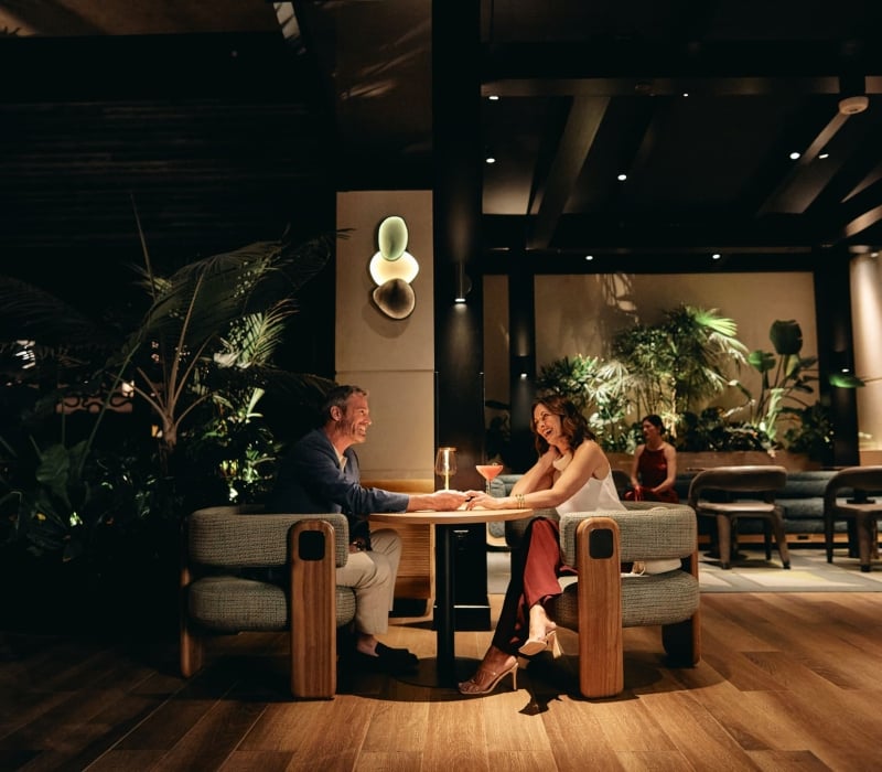 A smiling man and woman sit at a table, holding hands and talking over cocktails in a dimly lit, upscale lounge with wood floors and lush plants.