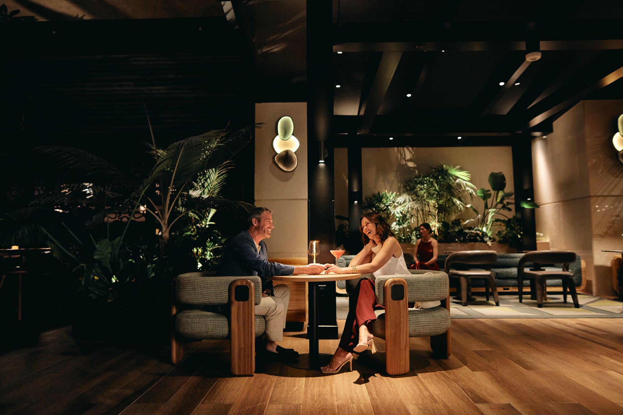 A smiling man and woman sit at a table, holding hands and talking over cocktails in a dimly lit, upscale lounge with wood floors and lush plants.
