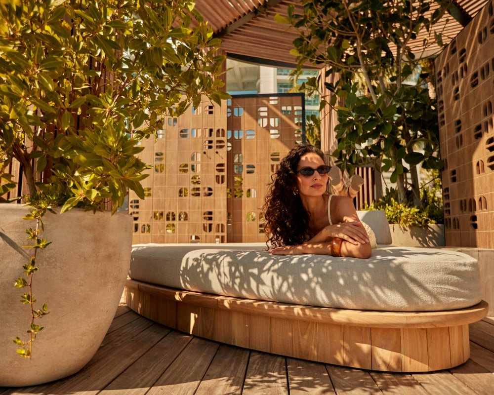 A woman with curly hair and sunglasses lounges on a cushioned daybed, resting her head on her hand. She is in a sunlit outdoor area with potted plants and decorative, perforated privacy screens.