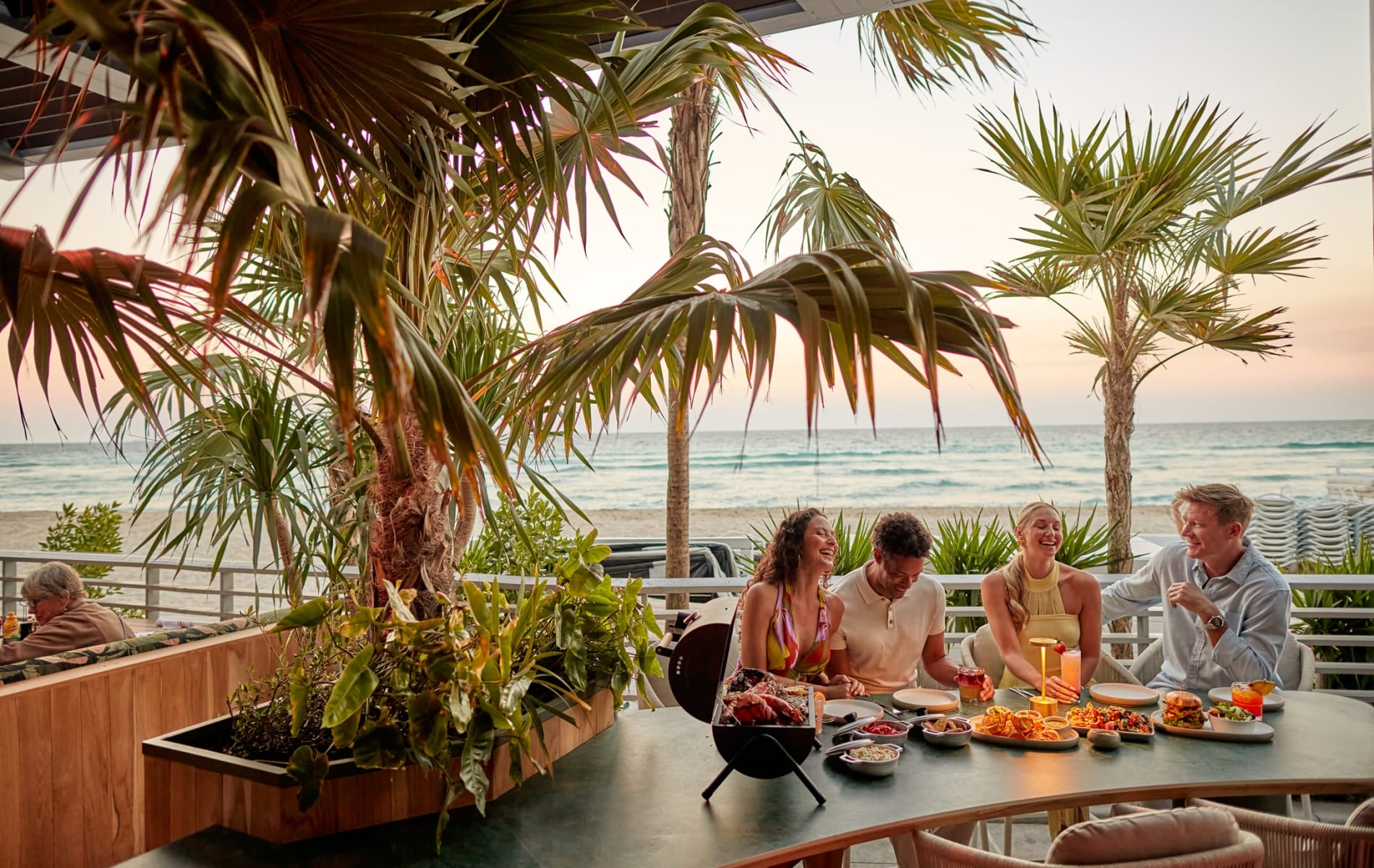 Four people laugh and share a meal with drinks on a patio table. Palm trees frame the scene, with an ocean beach visible at sunset.