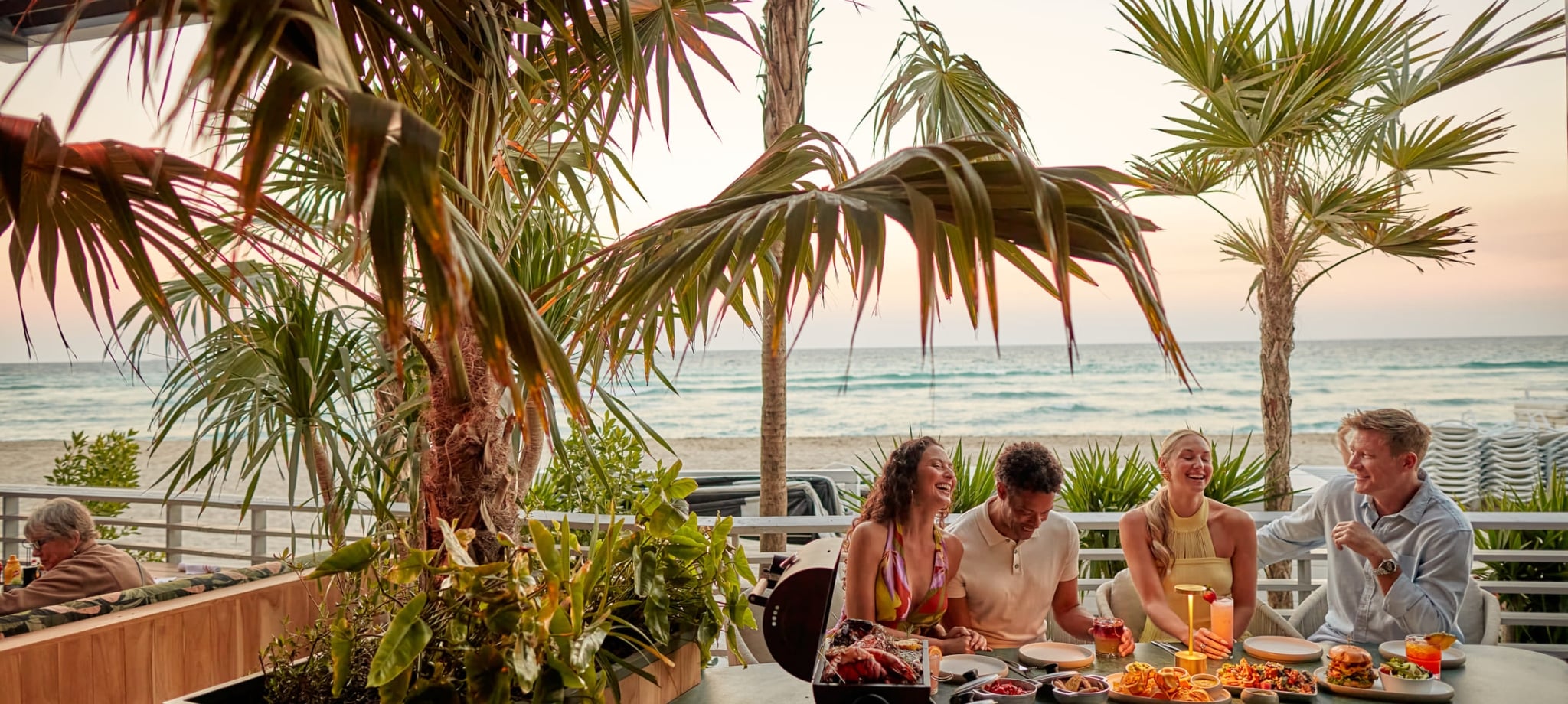Four people laugh and share a meal with drinks on a patio table. Palm trees frame the scene, with an ocean beach visible at sunset.