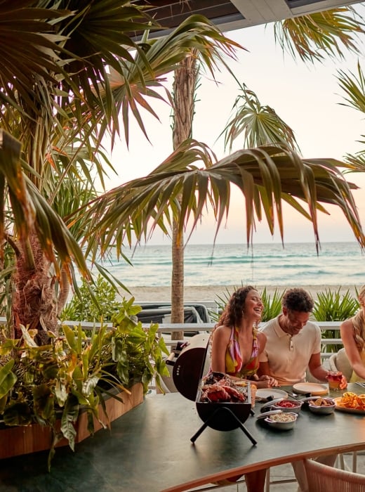 Four people laugh and share a meal with drinks on a patio table. Palm trees frame the scene, with an ocean beach visible at sunset.