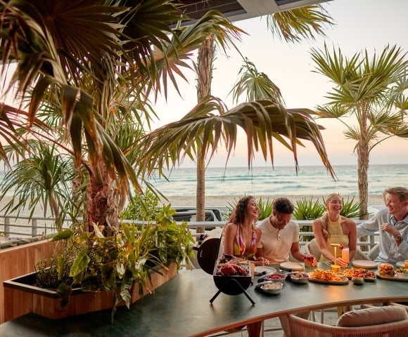Four people laugh and share a meal with drinks on a patio table. Palm trees frame the scene, with an ocean beach visible at sunset.
