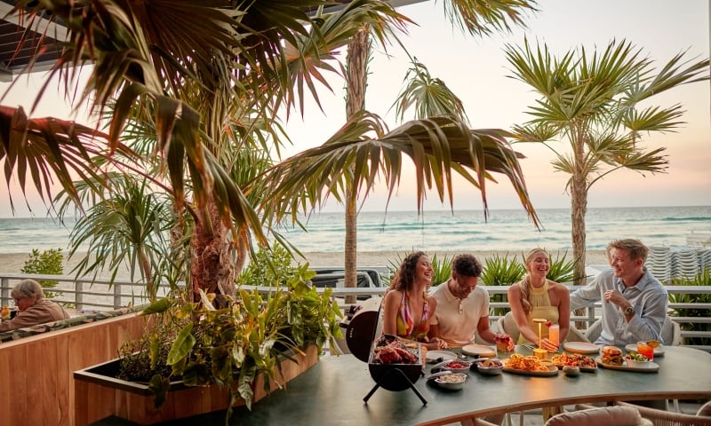Four people laugh and share a meal with drinks on a patio table. Palm trees frame the scene, with an ocean beach visible at sunset.