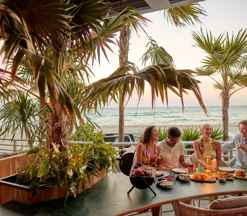 Four people laugh and share a meal with drinks on a patio table. Palm trees frame the scene, with an ocean beach visible at sunset.
