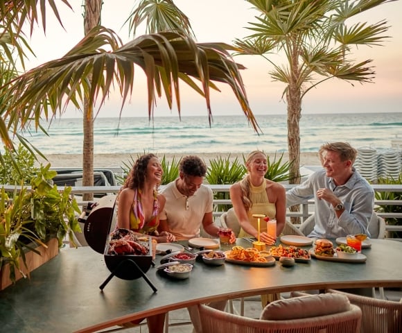 Four people laugh and share a meal with drinks on a patio table. Palm trees frame the scene, with an ocean beach visible at sunset.