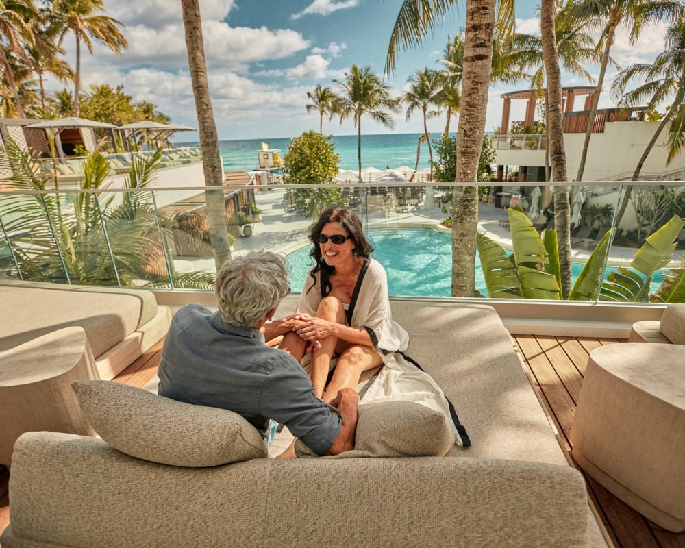 A couple sits and smiles on an outdoor lounge bed, overlooking a resort pool and the ocean with palm trees under a sunny sky.