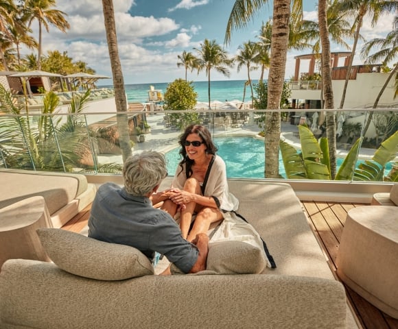A couple sits and smiles on an outdoor lounge bed, overlooking a resort pool and the ocean with palm trees under a sunny sky.