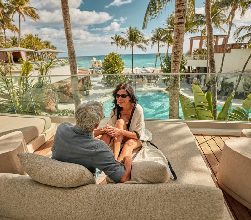 A couple sits and smiles on an outdoor lounge bed, overlooking a resort pool and the ocean with palm trees under a sunny sky.