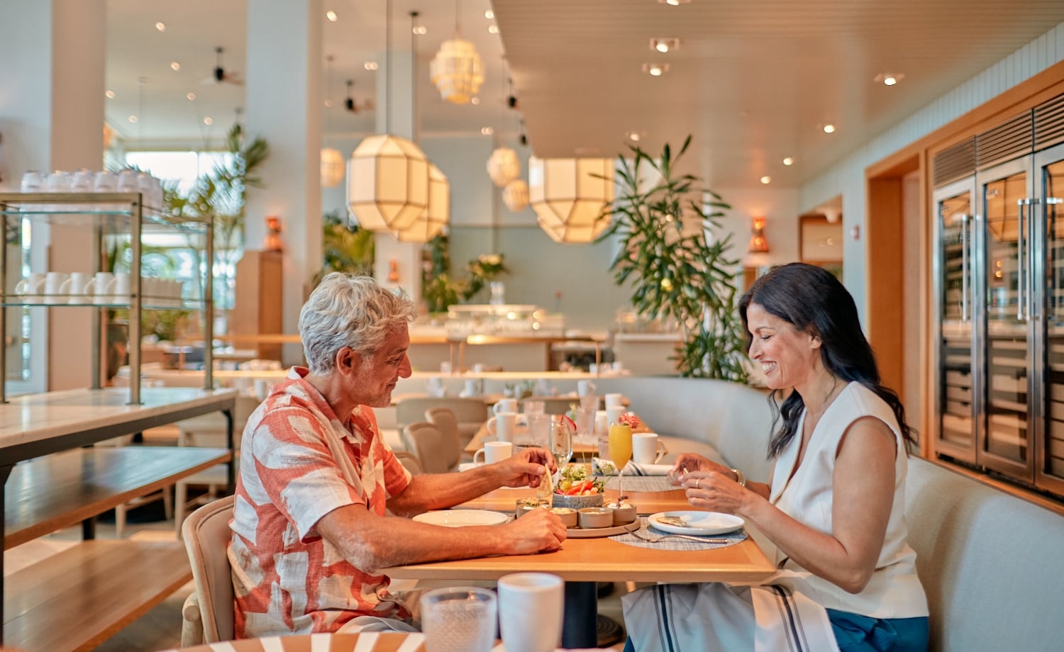 A man and woman happily eat small plates and drink beverages at a light wooden table. They are in a modern, well-lit restaurant with large geometric light fixtures and plants.