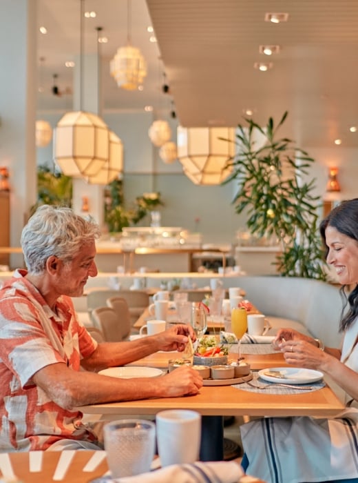 A man and woman happily eat small plates and drink beverages at a light wooden table. They are in a modern, well-lit restaurant with large geometric light fixtures and plants.