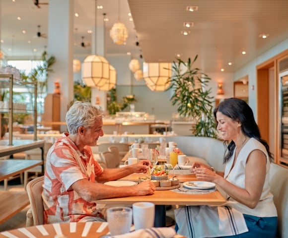 A man and woman happily eat small plates and drink beverages at a light wooden table. They are in a modern, well-lit restaurant with large geometric light fixtures and plants.