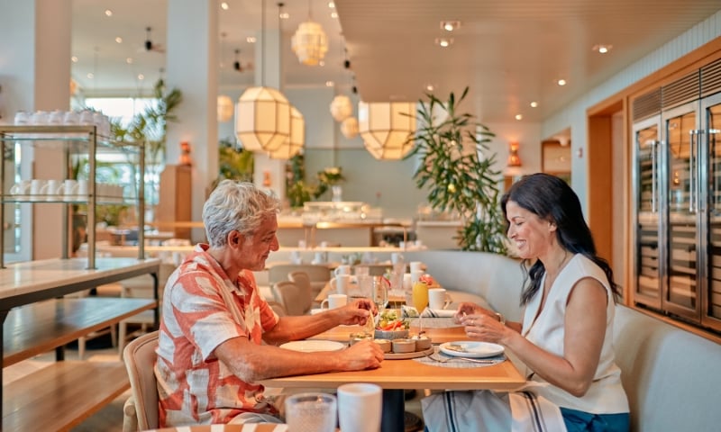A man and woman happily eat small plates and drink beverages at a light wooden table. They are in a modern, well-lit restaurant with large geometric light fixtures and plants.