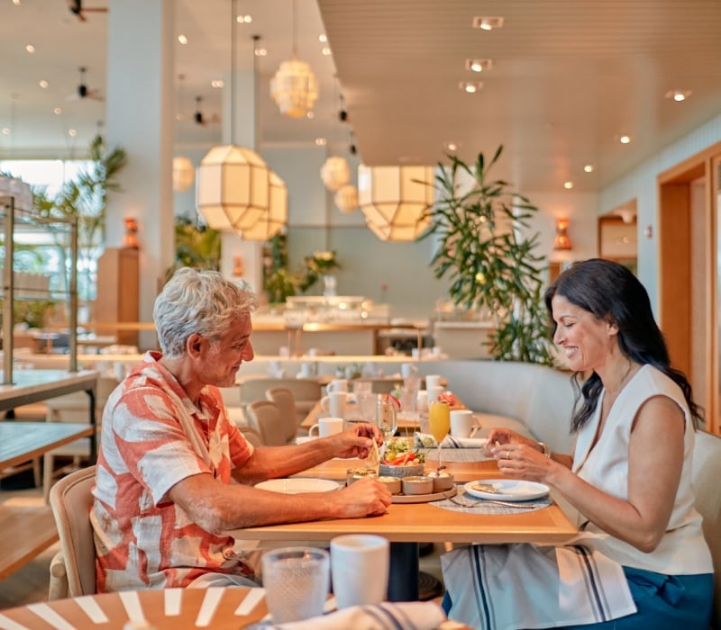 A man and woman happily eat small plates and drink beverages at a light wooden table. They are in a modern, well-lit restaurant with large geometric light fixtures and plants.