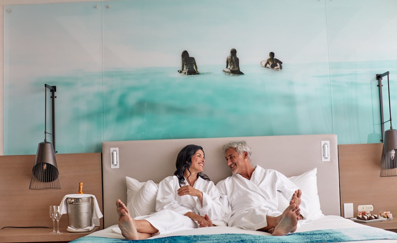 Smiling man and woman in white bathrobes sit barefoot on a bed, facing each other. Their hotel room features an ocean mural and champagne on a side table.