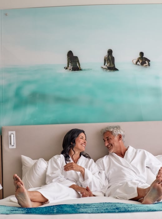 Smiling man and woman in white bathrobes sit barefoot on a bed, facing each other. Their hotel room features an ocean mural and champagne on a side table.