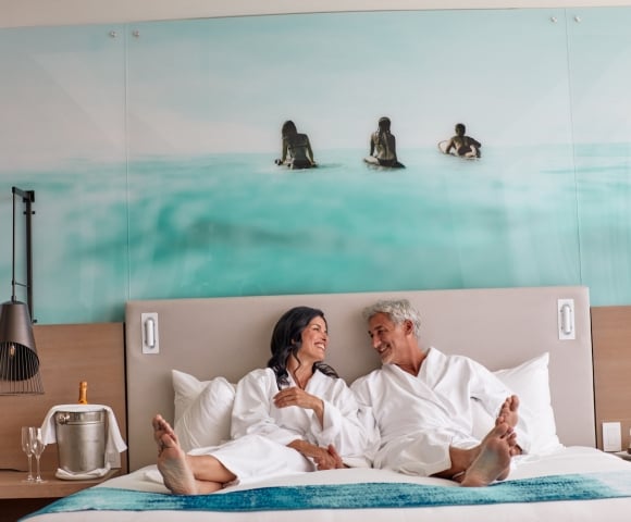 Smiling man and woman in white bathrobes sit barefoot on a bed, facing each other. Their hotel room features an ocean mural and champagne on a side table.