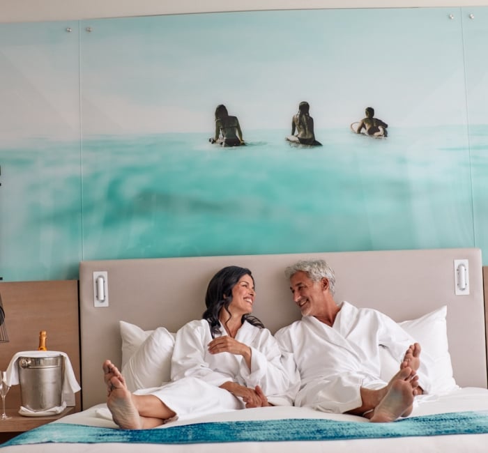 Smiling man and woman in white bathrobes sit barefoot on a bed, facing each other. Their hotel room features an ocean mural and champagne on a side table.