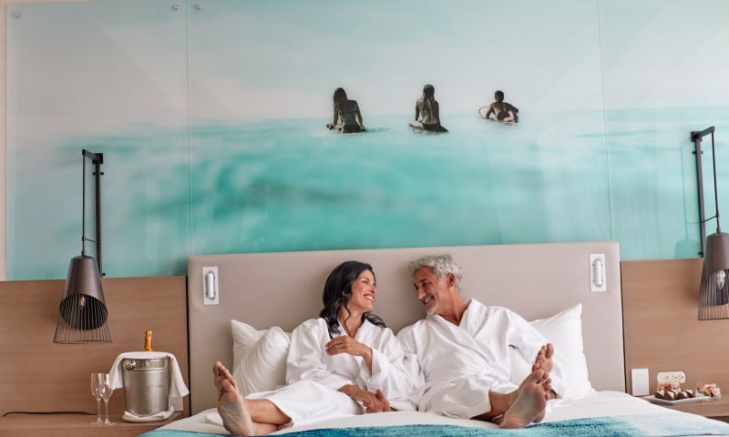 Smiling man and woman in white bathrobes sit barefoot on a bed, facing each other. Their hotel room features an ocean mural and champagne on a side table.