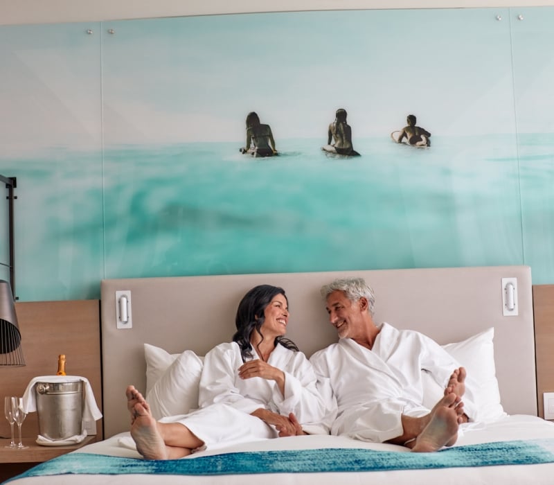 Smiling man and woman in white bathrobes sit barefoot on a bed, facing each other. Their hotel room features an ocean mural and champagne on a side table.