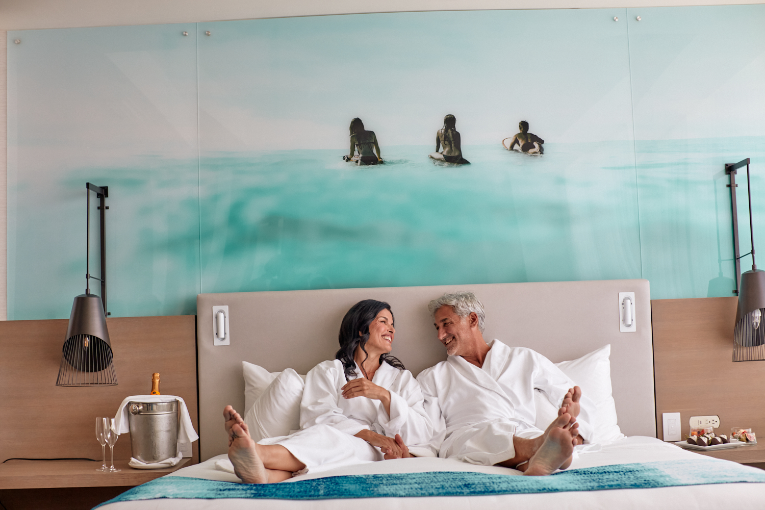 Smiling man and woman in white bathrobes sit barefoot on a bed, facing each other. Their hotel room features an ocean mural and champagne on a side table.