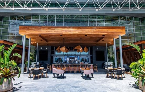 A stylish outdoor bar with seating under a wooden canopy, situated on a paved patio in front of a large glass building with an exposed steel truss roof.