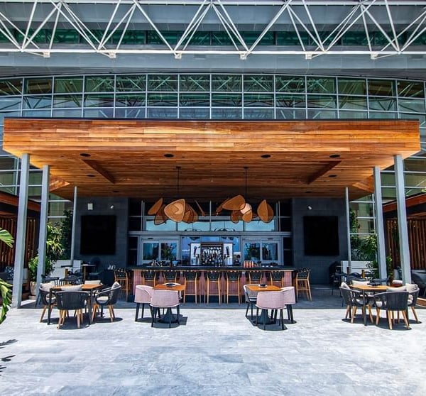 A stylish outdoor bar with seating under a wooden canopy, situated on a paved patio in front of a large glass building with an exposed steel truss roof.
