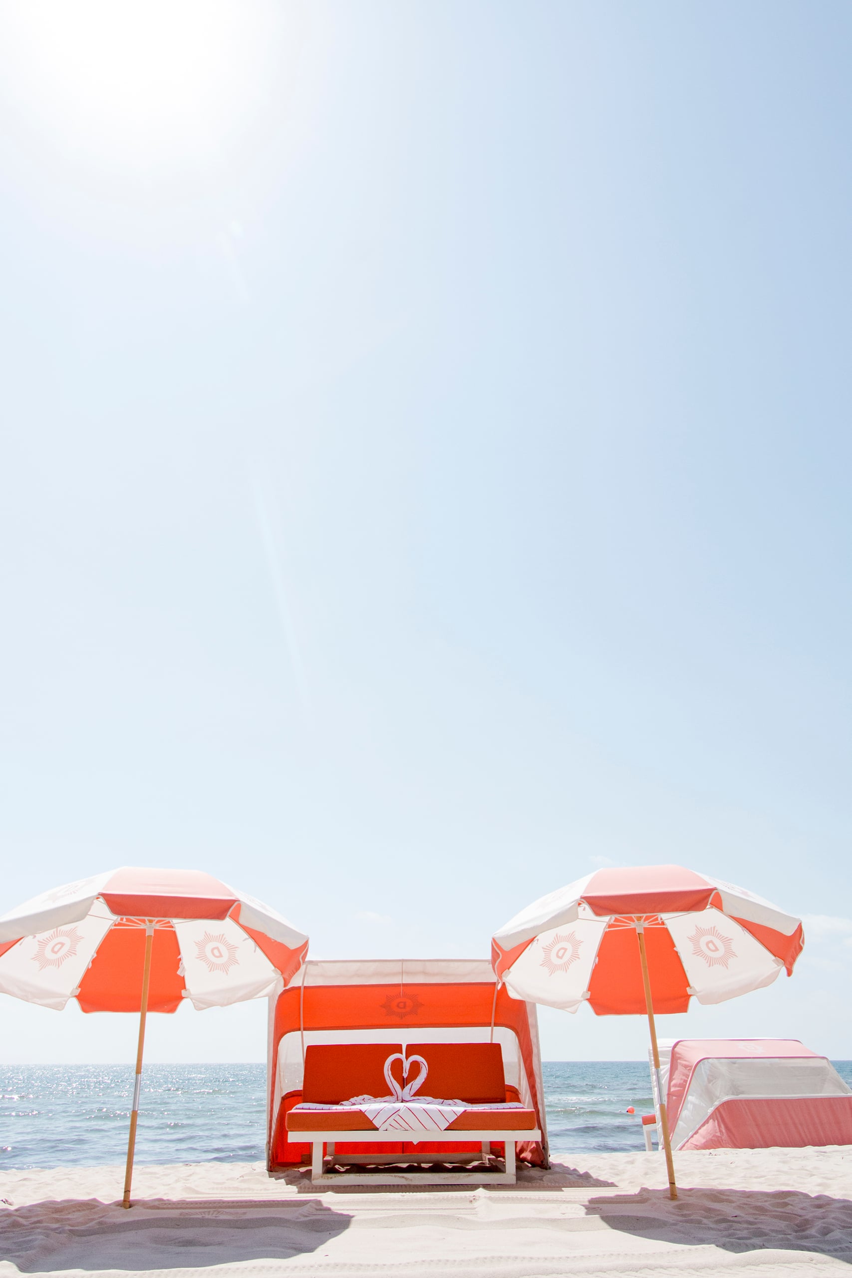 Orange-and-white striped beach cabana with swan towel art on a lounge bed, flanked by two umbrellas displaying a sun logo with 'D' inside, on a sandy beach by a sparkling ocean.