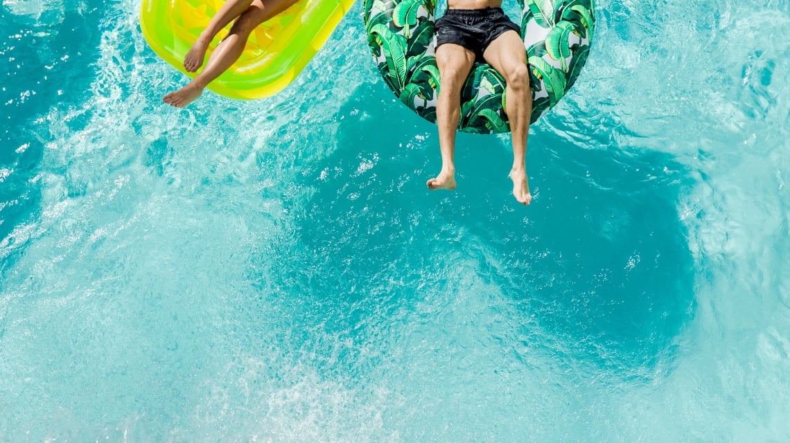 Two people float on distinct inflatable rafts—a yellow mat and a tropical leaf ring—in a clear, bright blue swimming pool, seen from above.