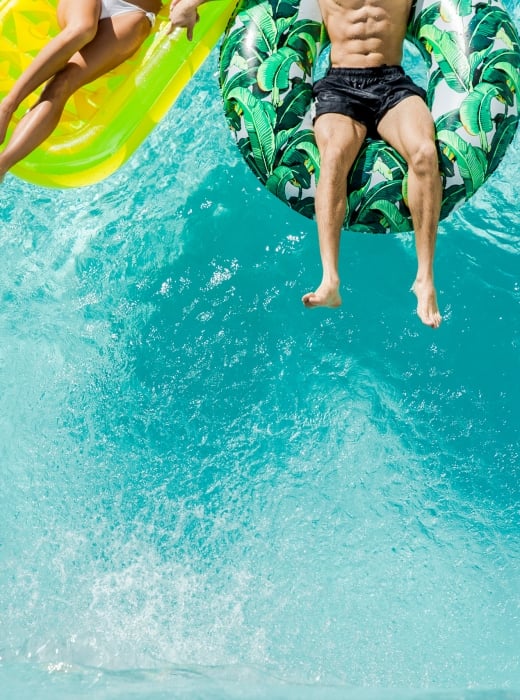 Two people float on distinct inflatable rafts—a yellow mat and a tropical leaf ring—in a clear, bright blue swimming pool, seen from above.