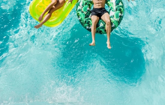 Two people float on distinct inflatable rafts—a yellow mat and a tropical leaf ring—in a clear, bright blue swimming pool, seen from above.