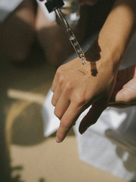 A hand receives a liquid drop from a glass dropper. "MUSE" is written on the hand, illuminated by warm natural light.