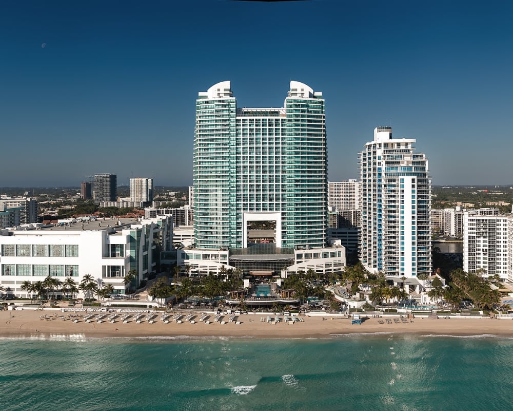 "DIPLOMAT" twin-towered building stands tall on a sandy beach by turquoise ocean water, surrounded by other hotels and a distant city skyline under a clear blue sky.