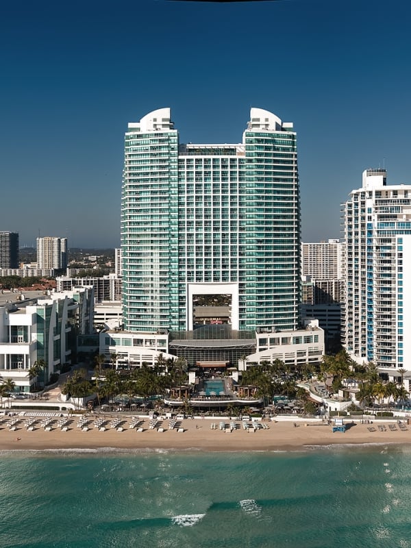 "DIPLOMAT" twin-towered building stands tall on a sandy beach by turquoise ocean water, surrounded by other hotels and a distant city skyline under a clear blue sky.
