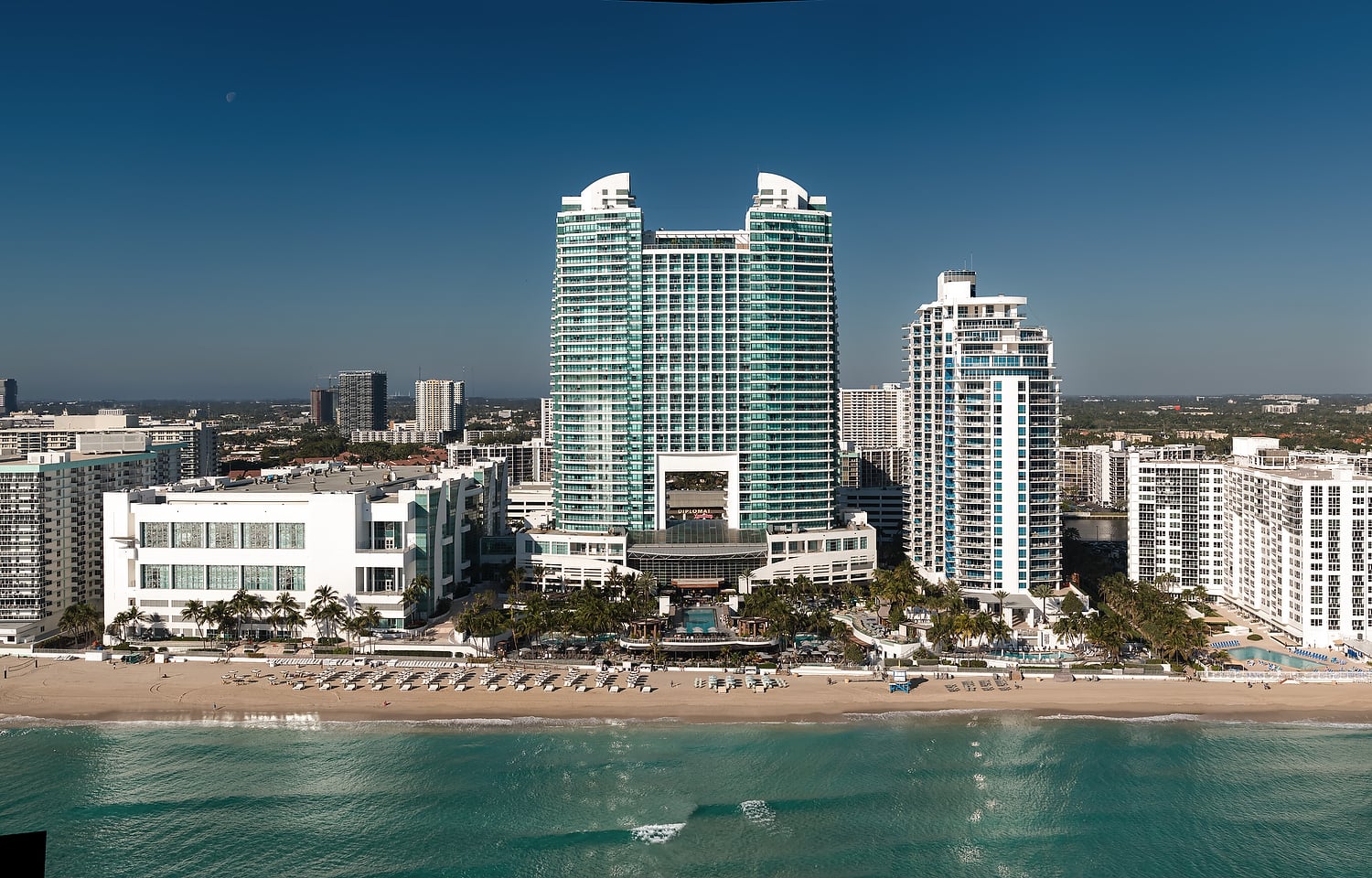 "DIPLOMAT" twin-towered building stands tall on a sandy beach by turquoise ocean water, surrounded by other hotels and a distant city skyline under a clear blue sky.