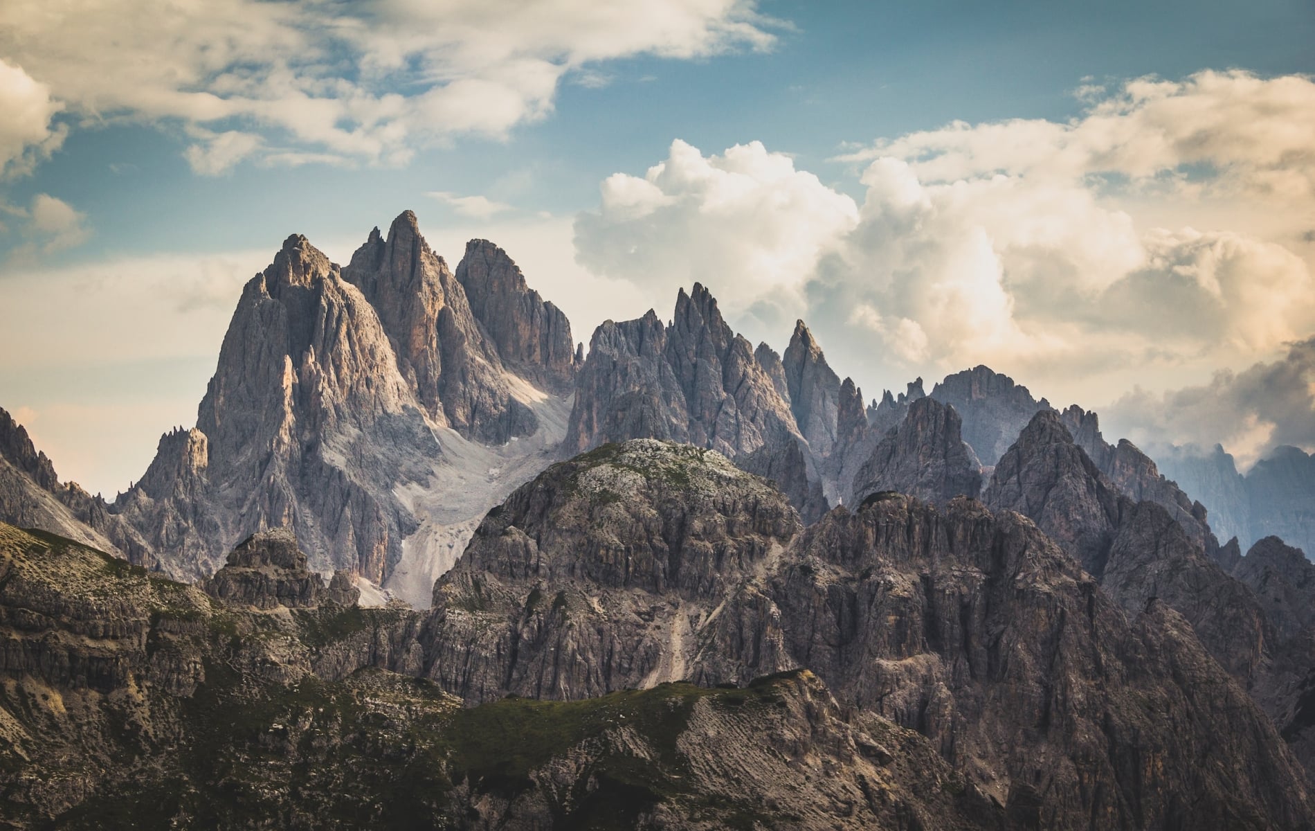 Jagged mountain peaks rise dramatically above rocky, green lower hills under a vast, cloudy sky, revealing distant valleys.
