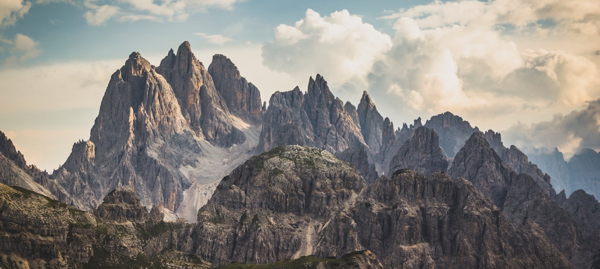 Jagged mountain peaks rise dramatically above rocky, green lower hills under a vast, cloudy sky, revealing distant valleys.